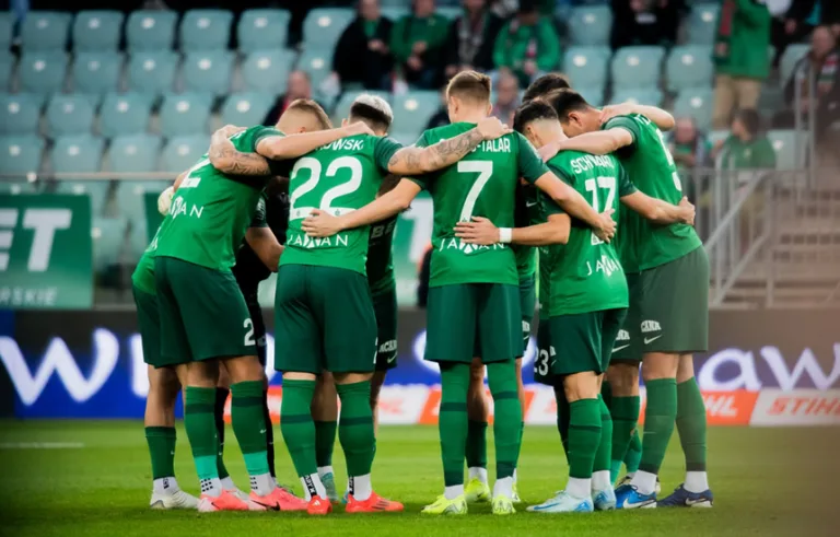 Piłkarze Śląska Wrocław stoją w kółku na murawie, motywując się przed meczem. W tle widać stadion, a atmosfera podkreśla zaangażowanie drużyny. Obrazek nawiązuje do wsparcia Fundacji Pociecha i kampanii - Zamień pączka na ciepły posiłek.
