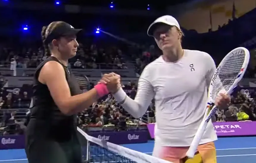 Iga Świątek podaje rękę Jelenie Ostapenko po zakończonym meczu w półfinale turnieju WTA 1000 w Doha. Obie tenisistki stoją przy siatce, Ostapenko z wyrazem triumfu, a Świątek z widocznym rozczarowaniem.