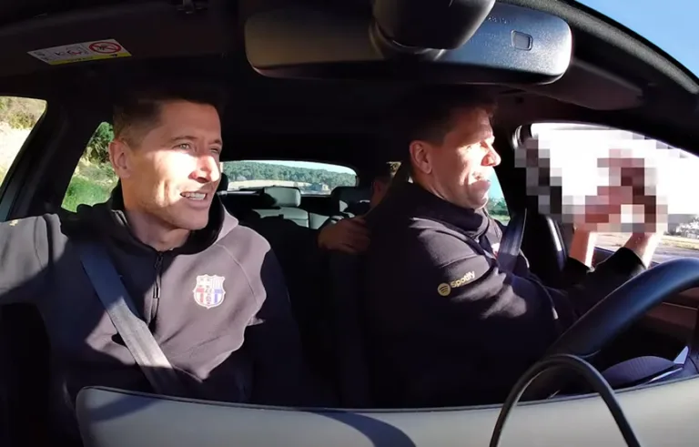 Wojciech Szczęsny siedzący za kierownicą samochodu marki Cupra, wykonujący żartobliwy gest przez szybę w stronę kolegów. Obok niego, na miejscu pasażera, Robert Lewandowski z uśmiechem obserwuje sytuację.