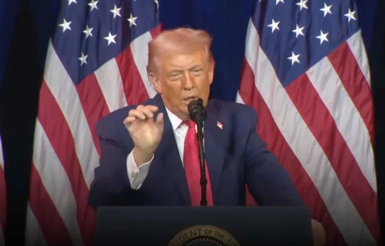 Donald Trump przemawia podczas wystąpienia publicznego, na tle amerykańskich flag, w kontekście napięć politycznych z Emmanuelem Macronem