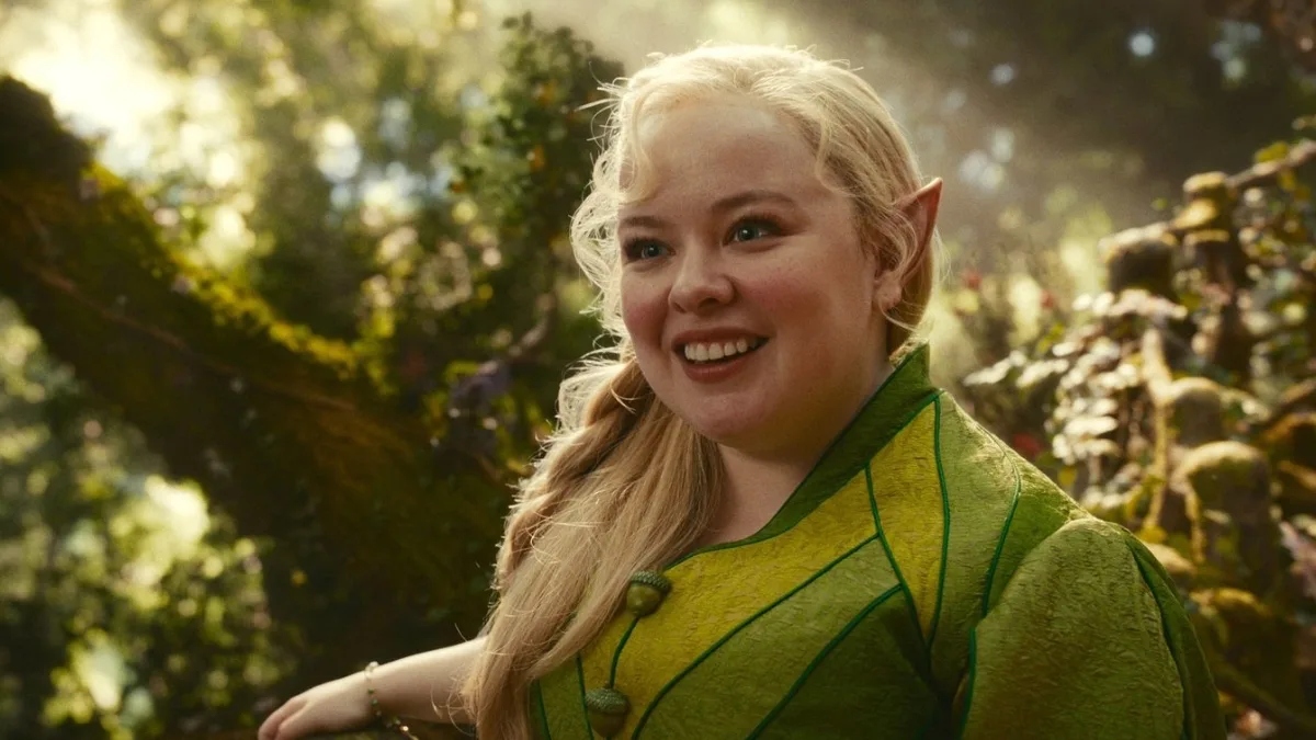 Uśmiechnięta blond dziewczyna z elfimi uszami w zielonym stroju stoi w magicznym lesie w scenie z filmu fantasy Drzewo Magii