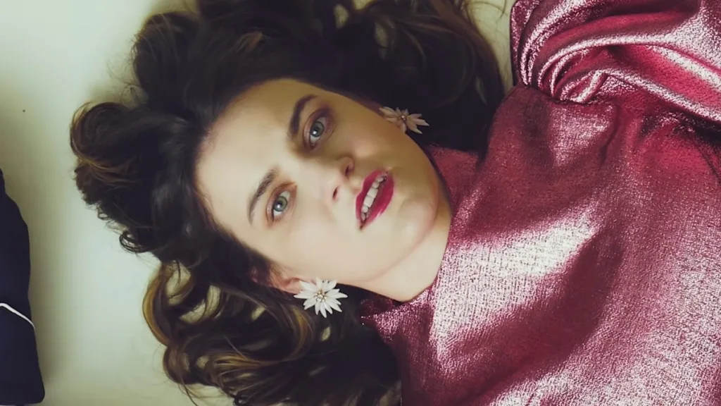 Hela Me Ry w kadrze z teledysku Butterfly M. Artystka w metalicznej różowej sukni i kwiatowych kolczykach leży na jasnym tle, patrząc intensywnie w kamerę.