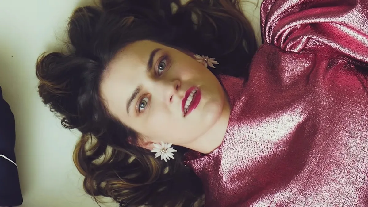 Hela Me Ry w kadrze z teledysku Butterfly M. Artystka w metalicznej różowej sukni i kwiatowych kolczykach leży na jasnym tle, patrząc intensywnie w kamerę.