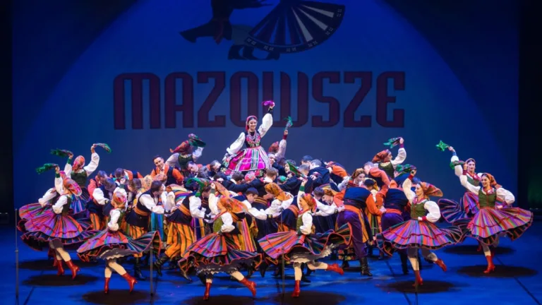 Artyści Zespół Pieśni i Tańca "Mazowsze" podczas barwnego występu na scenie, tancerze w tradycyjnych, kolorowych strojach ludowych wykonują dynamiczny układ choreograficzny, w tle widoczny napis Mazowsze.