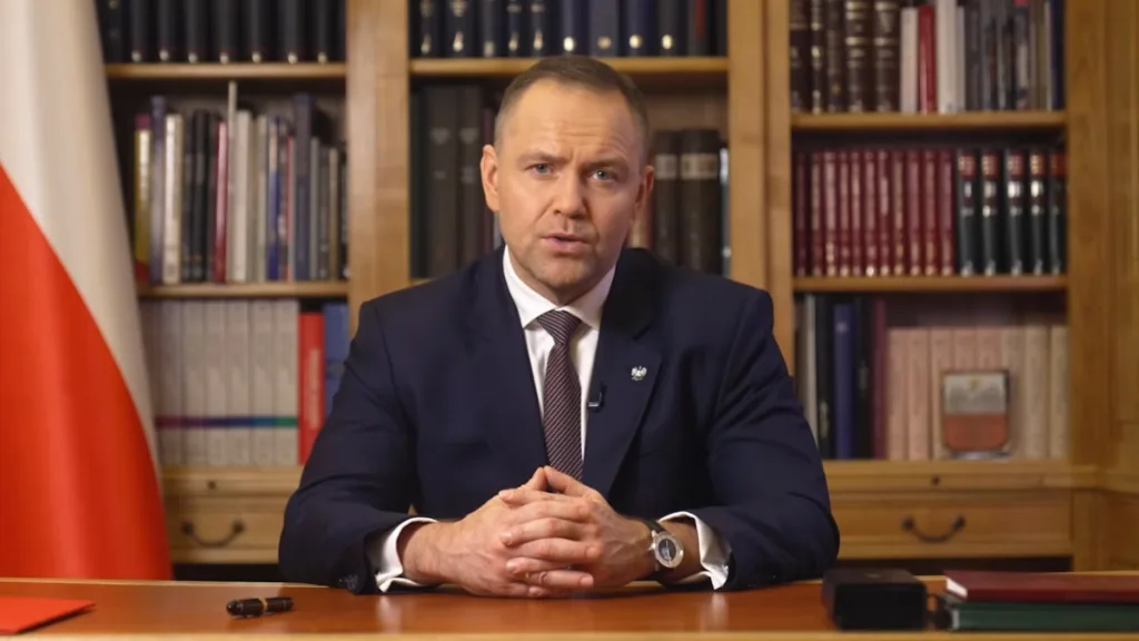 Karol Nawrocki wygłasza oficjalne wystąpienie w gabinecie, siedząc przy biurku na tle biblioteczki i flagi Polski.