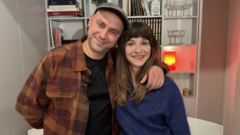 Damian Sikorski i Penelov stoją obok siebie w domowym wnętrzu, na tle półki z książkami i zapaloną lampką.