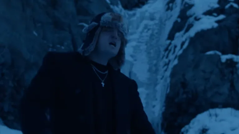 Guzior w zimowej scenerii przy zamarzniętym wodospadzie, w ciemnym płaszczu i futrzanej czapce w teledysku do "Przy sonecie"