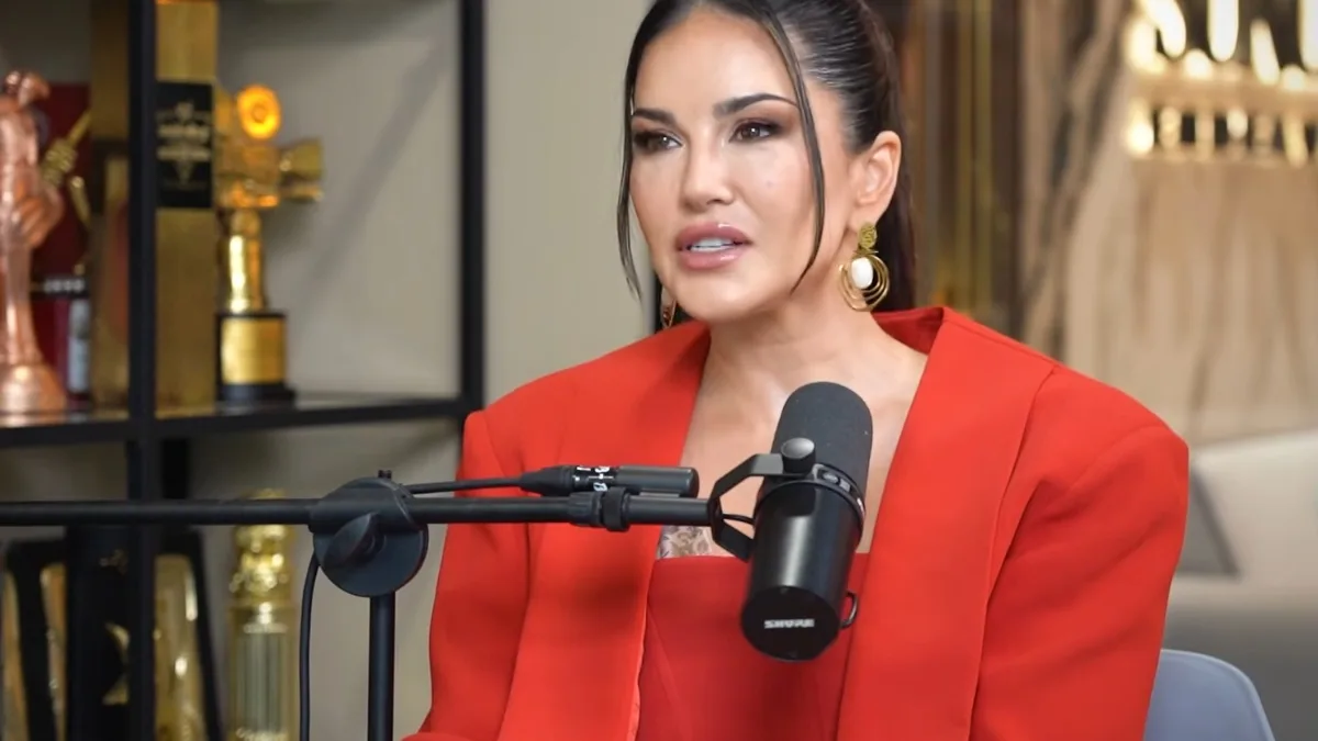 Sunny Leone w czerwonym garniturze podczas wywiadu, siedzi przed mikrofonem w studiu nagraniowym.