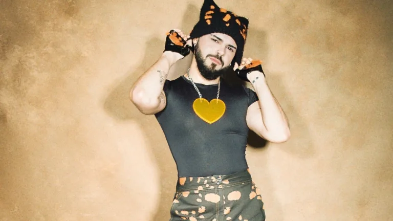 Akylas w czarnej dopasowanej koszulce i czapce z pomarańczowym wzorem pozuje na beżowym tle, trzymając dłonie przy głowie, promując singiel Ferto