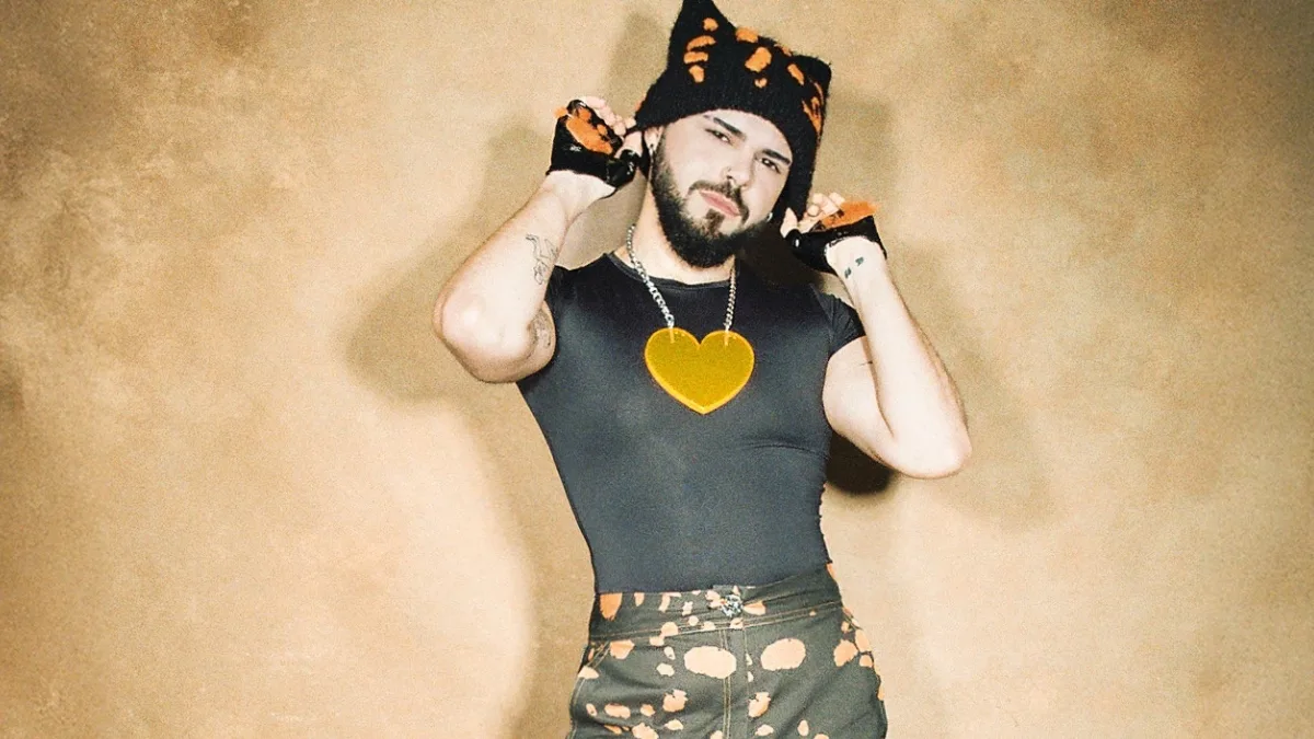 Akylas w czarnej dopasowanej koszulce i czapce z pomarańczowym wzorem pozuje na beżowym tle, trzymając dłonie przy głowie, promując singiel Ferto