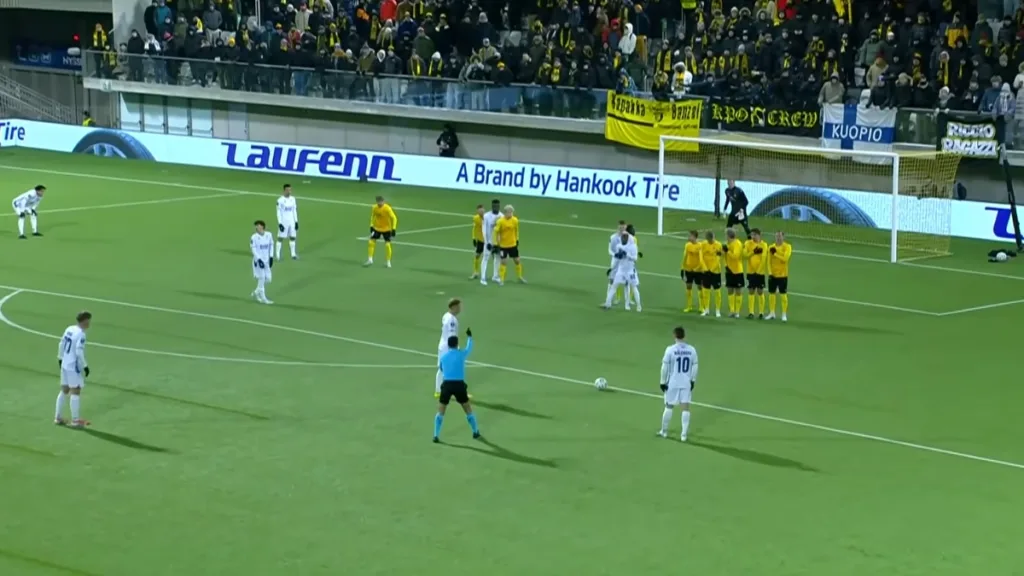 Rzut wolny dla Lecha Poznań w meczu z KuPS w 1/16 finału Ligi Konferencji - zawodnik w białym stroju przygotowuje się do strzału, przed nim mur piłkarzy KuPS w żółtych koszulkach.