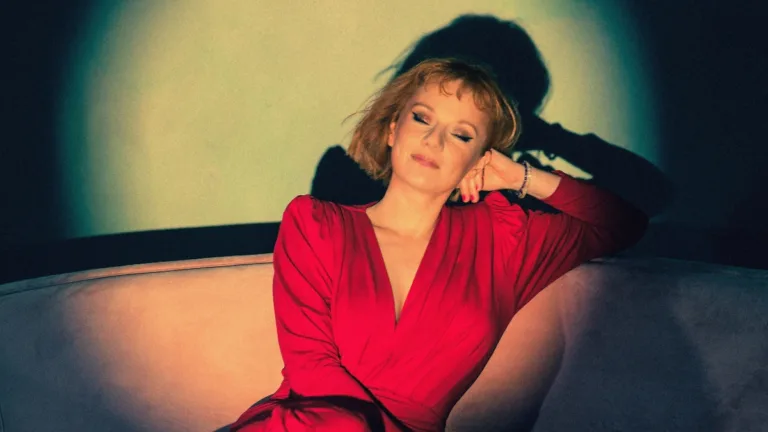 Ewelina Flinta w kadrze promującym album Mariposa siedzi na jasnej sofie w czerwonej sukience, z zamkniętymi oczami, w nastrojowym, ciepłym świetle.