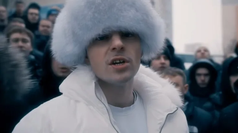 Lubin w dużej, futrzanej czapce i jasnej kurtce stoi na tle grupy ludzi w teledysku do utworu Prezydent. Kadr w chłodnej, miejskiej stylistyce.