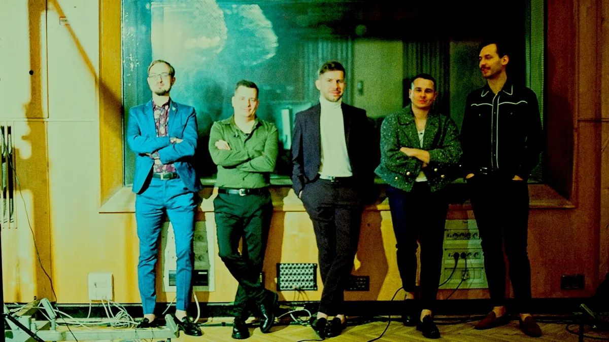 Martini Police stoi w studiu nagraniowym przy dużym oknie, oświetleni zielonkawym światłem; na podłodze widoczne są kable i elementy sprzętu audio.