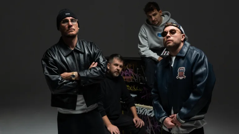 Czterech mężczyzn w stylizacjach hip-hopowych pozuje w studiu nagraniowym; dwóch w skórzanych kurtkach na pierwszym planie, w tle pianino z graffiti.