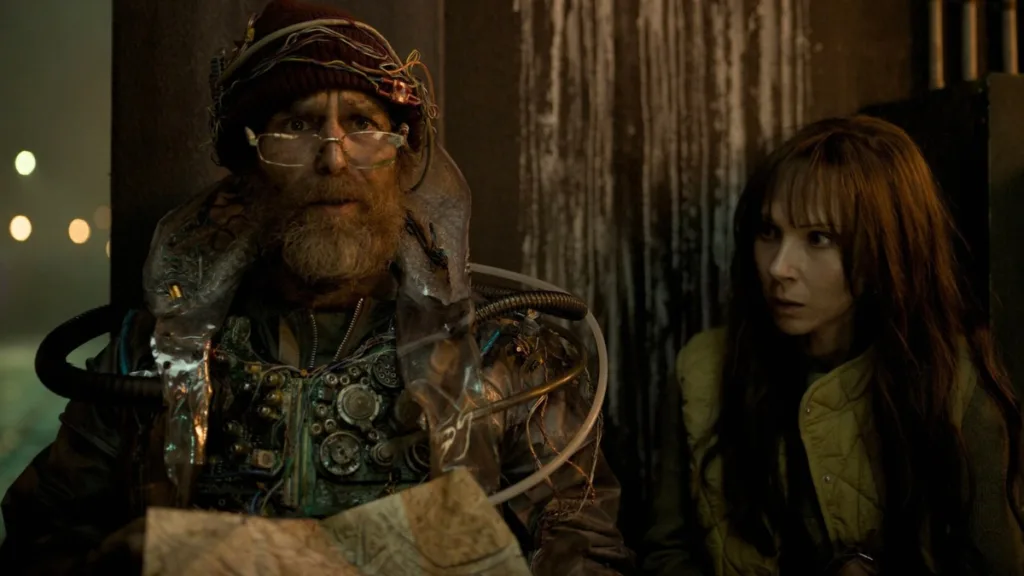 Kadr z filmu Baw się dobrze i przeżyj - bohater w technologicznej uprzęży i kobieta w napięciu, motyw AI kontra ludzkość.