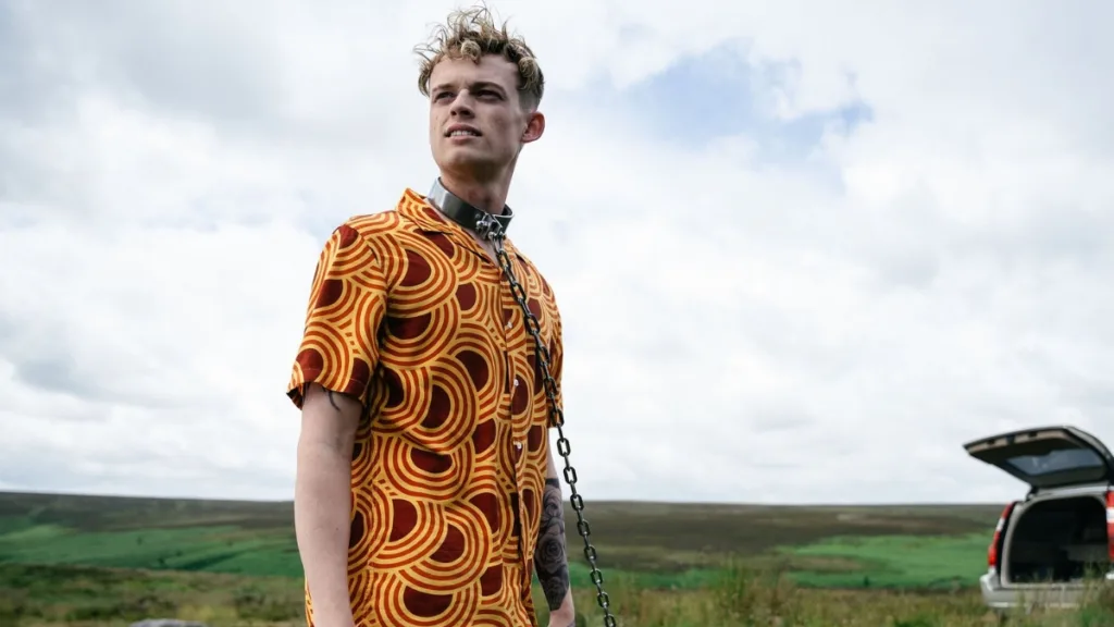 Młody mężczyzna w pomarańczowej koszuli stoi na otwartej przestrzeni z łańcuchem na szyi, w tle widać samochód i rozległy krajobraz.